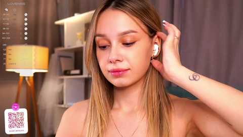 Media: Video of a light-skinned woman with long, straight blonde hair, wearing white wireless earbuds, and a tattoo on her right hand, in a cozy room with a beige lamp, shelves, and a gray curtain background.