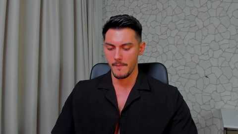 Media: Video of a clean-shaven man with short, dark hair, wearing a black shirt, sitting in a black chair in front of a textured gray wall with a white lamp visible in the background.