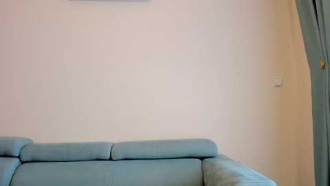 Media: Video of a minimalist living room with a light blue sectional sofa against a plain beige wall, featuring a small white switch and a partially visible curtain on the right.