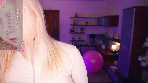 Media: Video of a blonde woman with long hair, viewed from behind, standing in a dimly lit living room with purple ambient lighting. The room has wooden furniture, shelves, and a pink exercise ball.