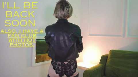 Media: Video of a woman with short blonde hair, wearing a black leather jacket and black lace panties, standing in a dimly lit room with green furniture and a mirror.