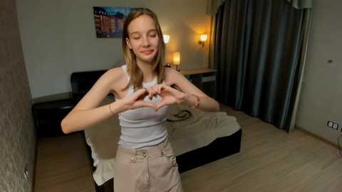 Media: Video of a slender, young Caucasian woman with fair skin and straight blonde hair, making a heart shape with her hands in a modern, minimalist bedroom. She wears a white tank top and beige shorts.