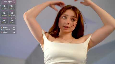 Media: Video of a slender, light-skinned woman with long brown hair, wearing a white tank top, performing a yoga pose indoors with a digital screen displaying weather information in the background.