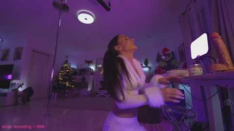 Media: Video of a woman in a white, fluffy, long-sleeve top and white pants, dancing with joy in a dimly lit, festive living room.