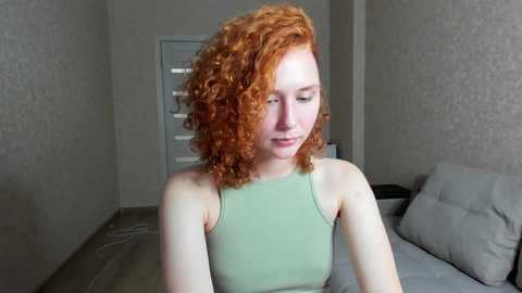 Media: Video of a fair-skinned woman with curly red hair, wearing a light green tank top, standing in a minimalist bedroom with beige walls and a gray couch.