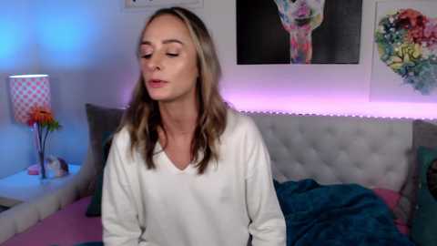 Media: Video of a blonde Caucasian woman in a white sweater, eyes closed, in a cozy bedroom with colorful floral art, pink lighting, and a teal bedspread.