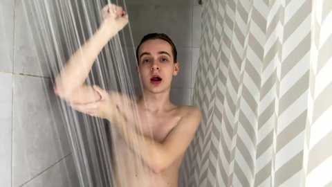 Media: A video of a slender, fair-skinned, nude man with short brown hair, standing in a shower with a white zigzag-patterned shower curtain. He holds a shower head in his right hand, water streaming down.