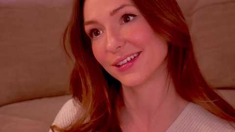 Media: Video of a smiling woman with fair skin and long, auburn hair, wearing a white knit sweater, seated against a beige couch background.