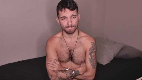 Media: Video of a shirtless, muscular, bearded Latino man with tattoos, wearing a necklace and a watch, sitting on a bed with a neutral background.