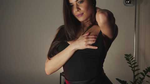 Media: Video of a woman with long dark hair, wearing a black top, touching her shoulder, against a dimly lit, beige wall with a plant in the background.