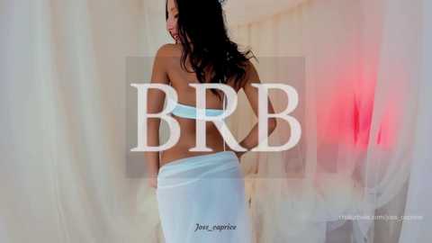 Media: Video of a dark-haired woman in a white towel, facing away, with \"BRR\" and \"Jane-negin\" text overlay. Background features sheer white curtains and a red light.