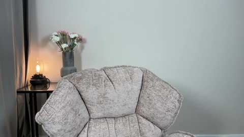 Media: A video of a beige, textured armchair with a cozy, dimly-lit lamp and a vase of pink and white flowers on a small, dark wooden table against a plain, light grey wall.