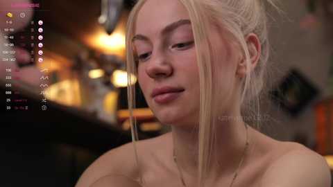 Media: Video of a fair-skinned young woman with platinum blonde hair in a messy bun, wearing a necklace, with a serene expression. Background shows blurred bar setting with warm lights.