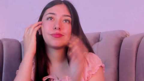 Media: Video of a young woman with long, straight black hair, wearing a pink, frilly top, sitting on a light gray, tufted sofa. She has fair skin, light makeup, and a soft smile.