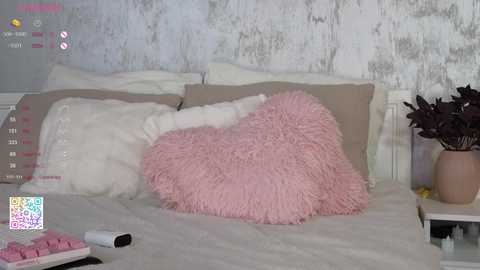 Media: Video of a cozy bedroom with a fluffy pink pillow, beige pillows, and a white throw on a bed. Background features a white shabby chic wall and a potted plant.