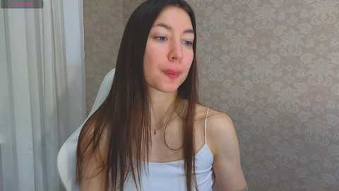 Media: Video of a young Caucasian woman with long, straight dark brown hair, wearing a white spaghetti-strap tank top, standing indoors with beige floral wallpaper background.