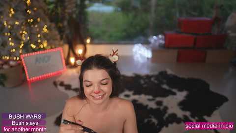 Media: A smiling young woman with fair skin, medium build, and dark hair adorned with antler headbands sits on a black-and-white cowhide rug in a festive, dimly-lit room. The background features a lit Christmas tree, red gift boxes, and glowing fairy lights. Text overlays mention \"BUSH WARS\" and \"social media big for another month.\