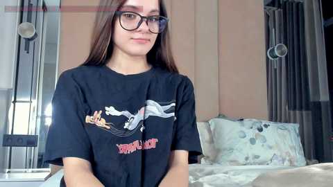 Media: Video of a young woman with long brown hair, wearing black-framed glasses and a black t-shirt with a colorful graphic, standing in a modern bedroom with a bed and white curtains.