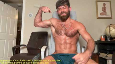 Media: Video of a muscular, shirtless man with a beard flexing his bicep, wearing green shorts, in a room with a closed door, chair, and framed artwork.