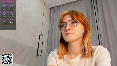 Media: A video of a fair-skinned woman with shoulder-length auburn hair, wearing round glasses and a white top, standing in a modern room with grey curtains. A digital display showing weather data is visible in the background.