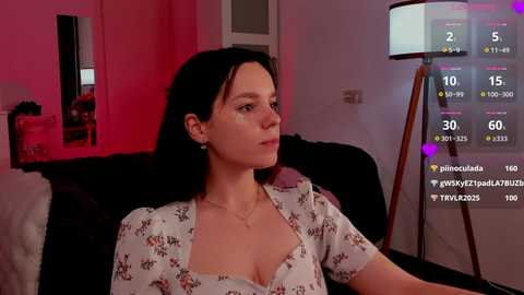 Media: Video of a pale-skinned woman with dark hair in a floral blouse, seated on a dark couch in a dimly lit room with a pinkish hue. A TV screen shows virtual reality gameplay statistics.