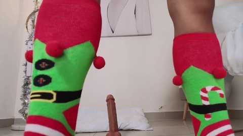 Media: Video of a person wearing festive Christmas-themed leggings with large red and green patches, standing in a bedroom with a white bed, a floor lamp, and a framed abstract artwork.