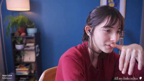 Media: Video of a young woman with dark hair, red sweater, and headphones, sitting pensively in a blue-walled room with a bookshelf, potted plant, and lamp.