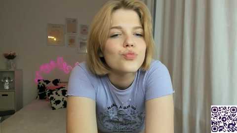 Video of a young blonde woman making a pouty face, wearing a light blue T-shirt with a graphic design, in a cozy, dimly lit bedroom with cow-print bedding, pink fairy lights, and a QR code watermark.