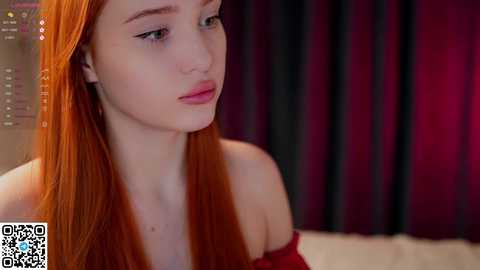 Media: Video of a young, fair-skinned woman with long, straight red hair, wearing a red off-shoulder top, looking thoughtful against a dark curtain background.