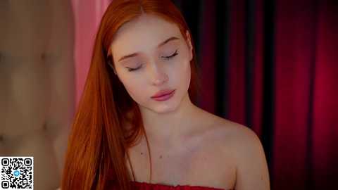 Media: Video of a young, fair-skinned woman with long, straight red hair, wearing a strapless, off-the-shoulder red dress. She has a serene expression, eyes closed, against a backdrop of dark curtains.