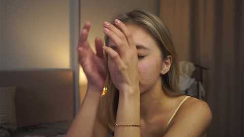 Media: Video of a young woman with light skin and blonde hair, covering her face with her hands, wearing a gold necklace and bracelet, in a dimly lit room with beige walls and a bed in the background.