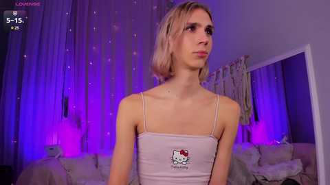 Media: Video of a slender, androgynous person with short blonde hair wearing a white tank top with a Hello Kitty logo, standing in a dimly lit room with purple lighting and curtains.