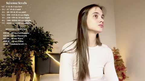 Media: Video of a young Caucasian woman with long brown hair, wearing a white sweater, standing in a room with beige walls and Christmas decorations.