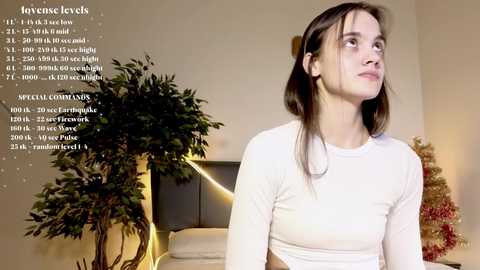 Media: Video of a young woman with fair skin and long brown hair, wearing a white long-sleeve top, standing indoors next to a large potted plant and a decorated Christmas tree.