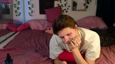 Video of a young man in a white t-shirt, lying on a bed with red and pink bedding, holding a bottle of beer, in a dimly lit bedroom.