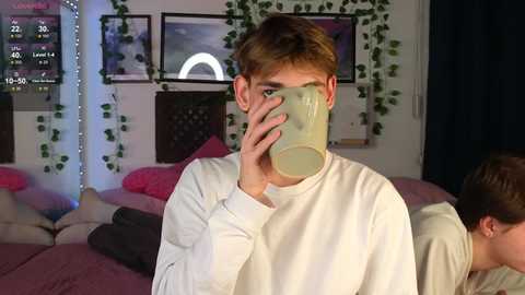 Video of a young man with light brown hair drinking from a green cup in a dimly lit, cozy bedroom with hanging vines, pink bedding, and a digital clock on the wall.