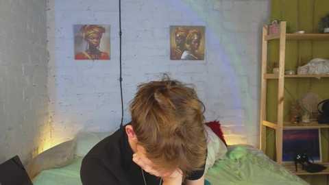 Media: Video of a person with short brown hair, wearing a black top, sitting on a bed with green sheets, looking at a white brick wall adorned with two paintings of Indian gods. A wooden shelf and a green wall are visible in the background.