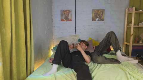 Video of a young man with fair skin and short brown hair lying on a bed, wearing a black shirt and gray pants, reading a book, in a cozy, brightly lit room with green walls and a wooden bookshelf.