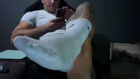 Media: Video of a muscular man with short dark hair, wearing white socks with black cartoon faces on them, sitting in a black chair, holding a smartphone, with a dimly lit room in the background.