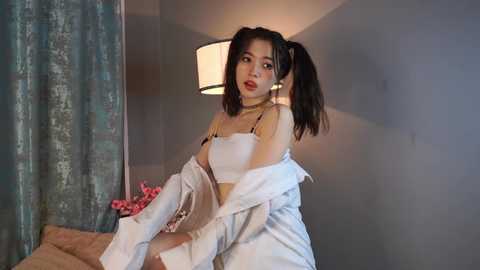 Media: Video of an Asian woman with pigtails, wearing a white crop top and matching robe, sitting on a bed with a pink floral pillow.