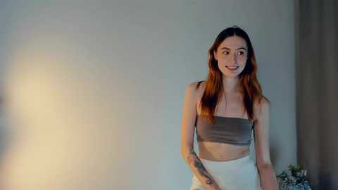 Media: Video of a slender, light-skinned woman with long, wavy auburn hair, wearing a grey bandeau top and white pants, standing against a plain beige wall, smiling softly.