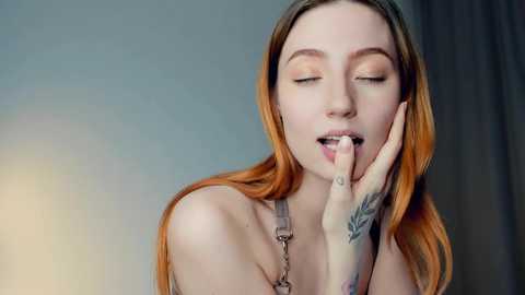 Media: Video of a fair-skinned woman with long, vibrant orange hair, wearing a grey strapless top, touching her lips with her hand. Her eyes are closed, and she has a tattoo on her left hand. Background is a blurred, neutral gradient.