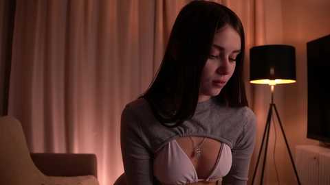 Media: Video of a young woman with straight dark hair, wearing a grey cropped top revealing a pink bra, in a dimly lit room with beige curtains and a black lamp.