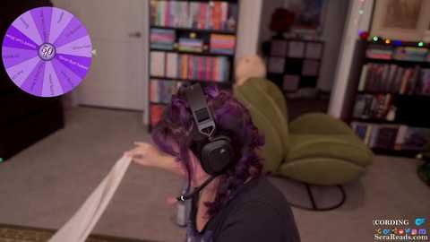 Video of a person with purple hair, wearing a black headset, lying on a beige carpet in a room with a green armchair, bookshelves, and a purple vinyl record displayed.