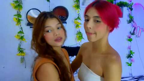 Media: Video of two young women with tan skin, one with long brown hair and orange top, the other with short red hair and white strapless top, smiling in a brightly lit room with yellow flower garlands and a round mirror on a white wall.