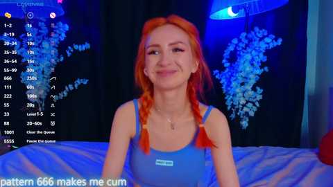 Media: Video of a smiling, fair-skinned woman with vibrant red braided hair, wearing a blue tank top, against a dark, blue-lit background.