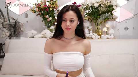 Media: Video of a young woman with long dark hair, wearing a white off-shoulder crop top and white pants, seated in a modern, minimalistic room with white walls, floral arrangements, and a white couch.