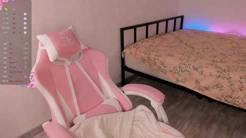 Media: Video of a pink gaming chair with a white and black headrest in a minimalist bedroom with a bed covered in a beige floral duvet, featuring a soft light blue glow from the floor.