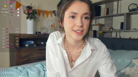 Media: Video of a young woman with fair skin, brown hair, and light makeup, wearing a white shirt and necklace, smiling in a cozy bedroom with a wooden dresser and bed.