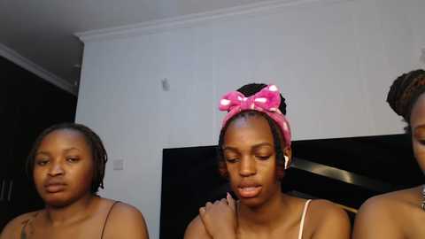 Media: Video of three young Black women with medium skin tones, wearing spaghetti straps, sitting in a living room. One wears a pink headband with polka dots, while another has a tattoo on her shoulder.